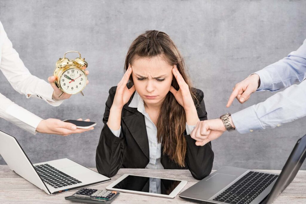 A woman sits at her desk, head in her hands, feeling overwhelmed by an endless list of tasks that are all causes of stress.