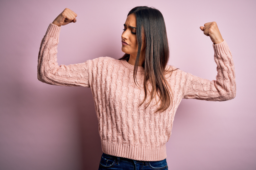 A woman in a pink sweater confidently flexing her arms to highlight the result of muscle and strength training.