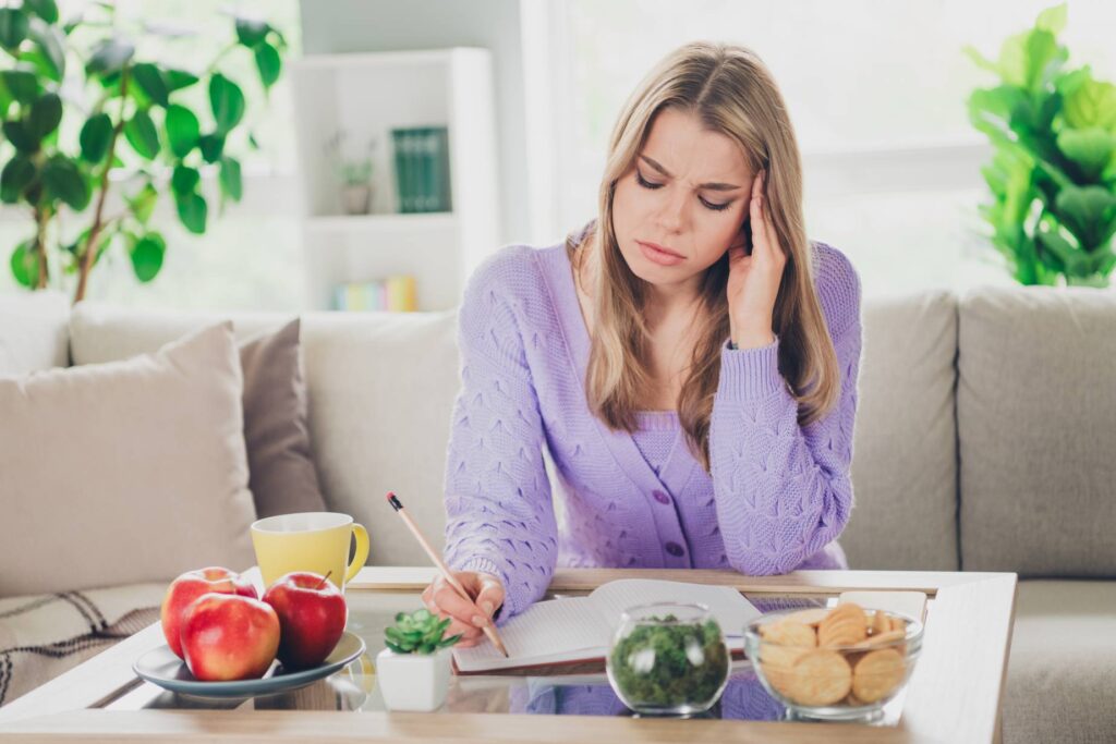 A woman sitting at a table with a pained expression, holding her head. Cookies and snacks surround her, suggesting she might get headaches after eating them, possibly due to food intolerances.